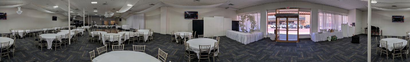 A wide banquet room set with round tables in white linens and gold chairs, with broad aisles between them. Glass double doors at the center of the far wall open at ground level onto the sidewalk and parking lot. Skirted tables line the window wall, and screens are mounted on the side walls.