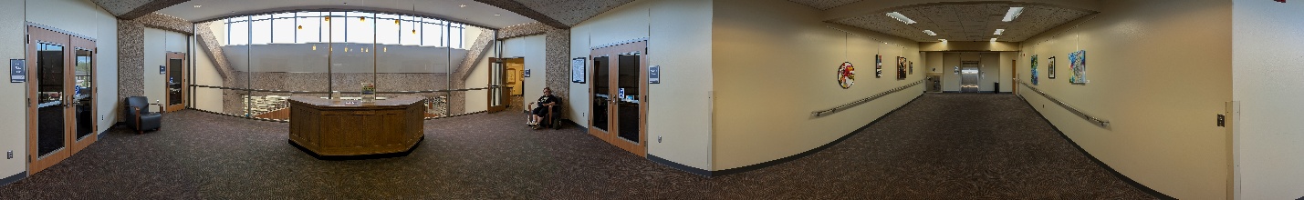 The library's second floor. Wood double doors to the meeting rooms open off a carpeted landing with an octagonal information desk, where a railing overlooks the main floor through tall arched windows. To the right, a corridor with handrails along both walls leads past hanging artwork to the elevator at the far end.
