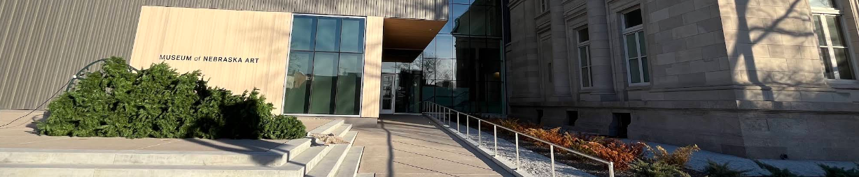 The museum entrance, set in the glass-walled gap between the modern limestone addition on the left and the historic stone building on the right. A railed ramp rises from the right side of the plaza to the door; wide steps approach from the left.